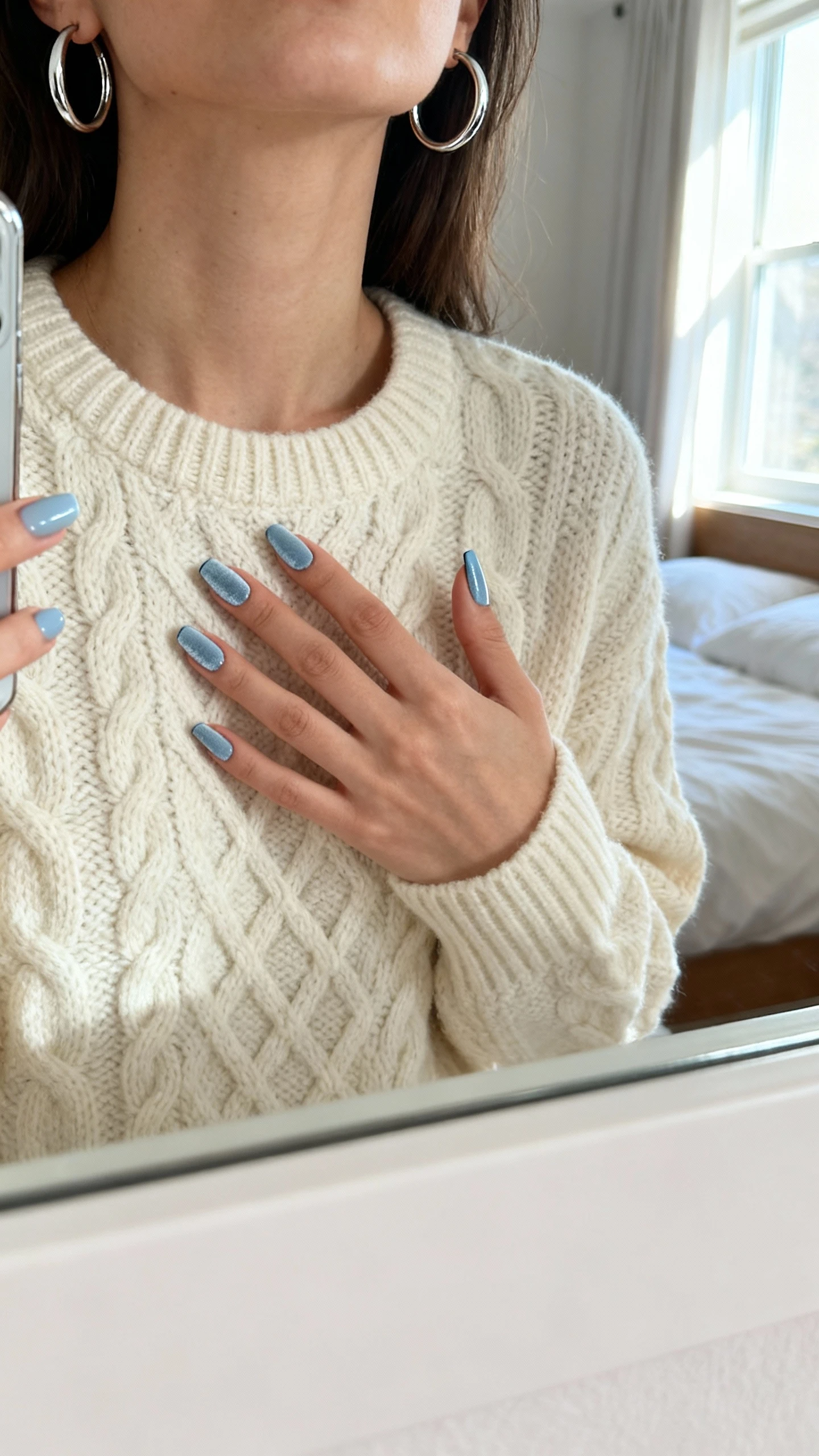 iPhone mirror selfie of a woman showing velvet-finish cat-eye nail polish in a soft icy blue that looks like cashmere, wearing a cream cable-knit sweater and silver hoop earrings, face not visible, cropped at neck, bedroom mirror with morning window light, iPhone photo quality.