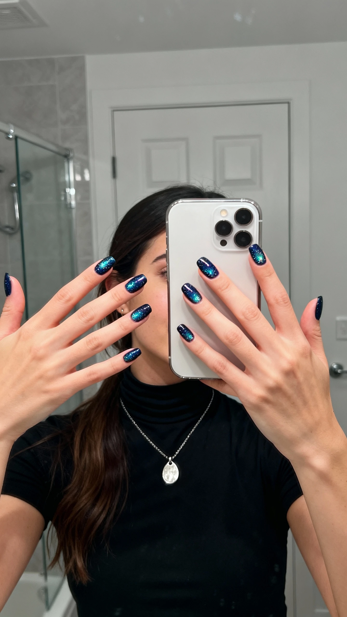 iPhone mirror selfie of a woman with galaxy nails (deep navy base with micro sparkle like starlit skies), wearing a black mock-neck top and silver pendant necklace, face not visible, bathroom mirror, cool indoor lighting, iPhone photo quality.