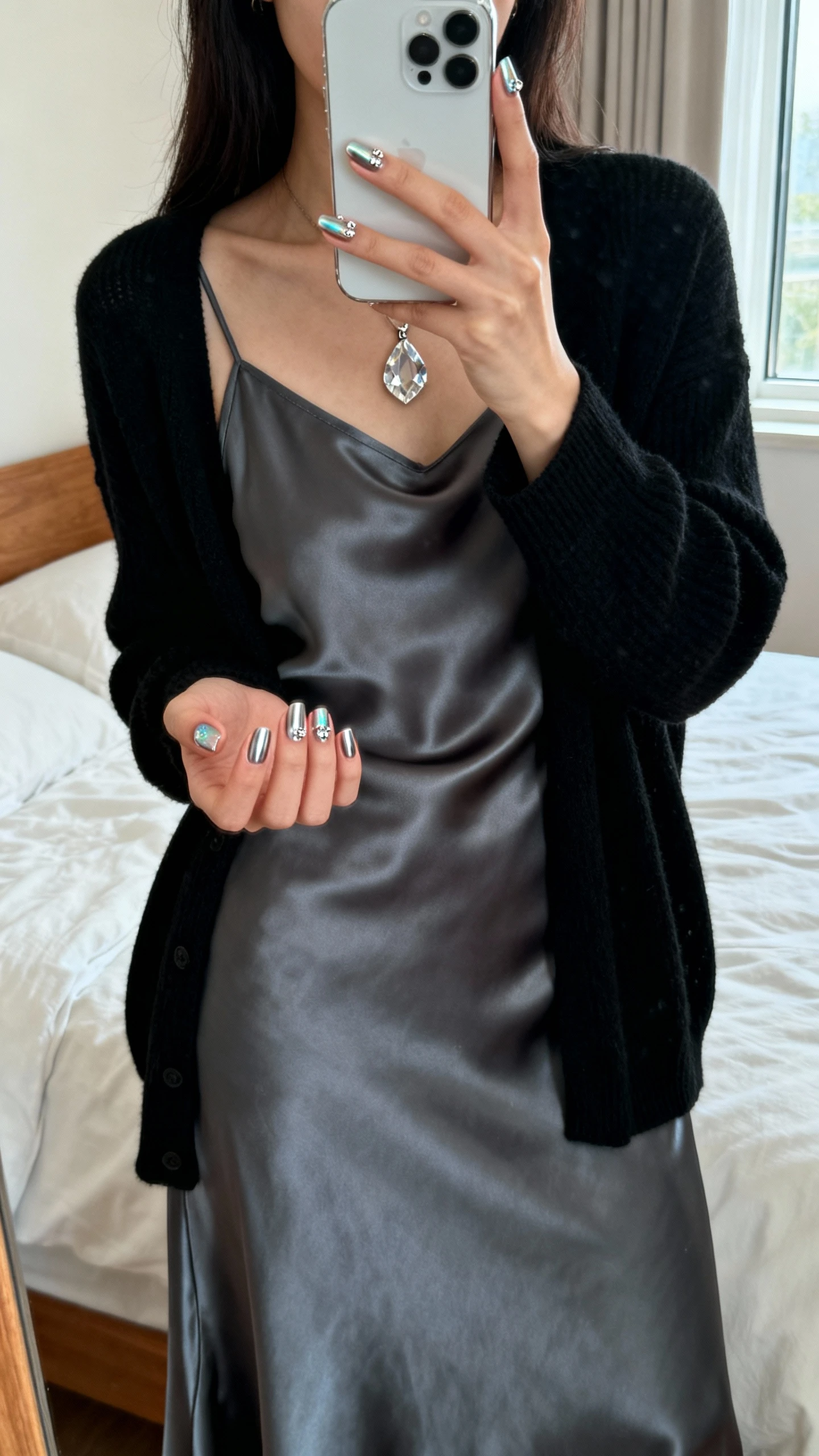iPhone mirror selfie of a woman with gemstone aura nails (subtle chrome base with tiny crystal accents), wearing a charcoal satin slip dress layered with a black cardigan and a crystal pendant, face not visible, bedroom mirror, natural window light, iPhone photo quality.