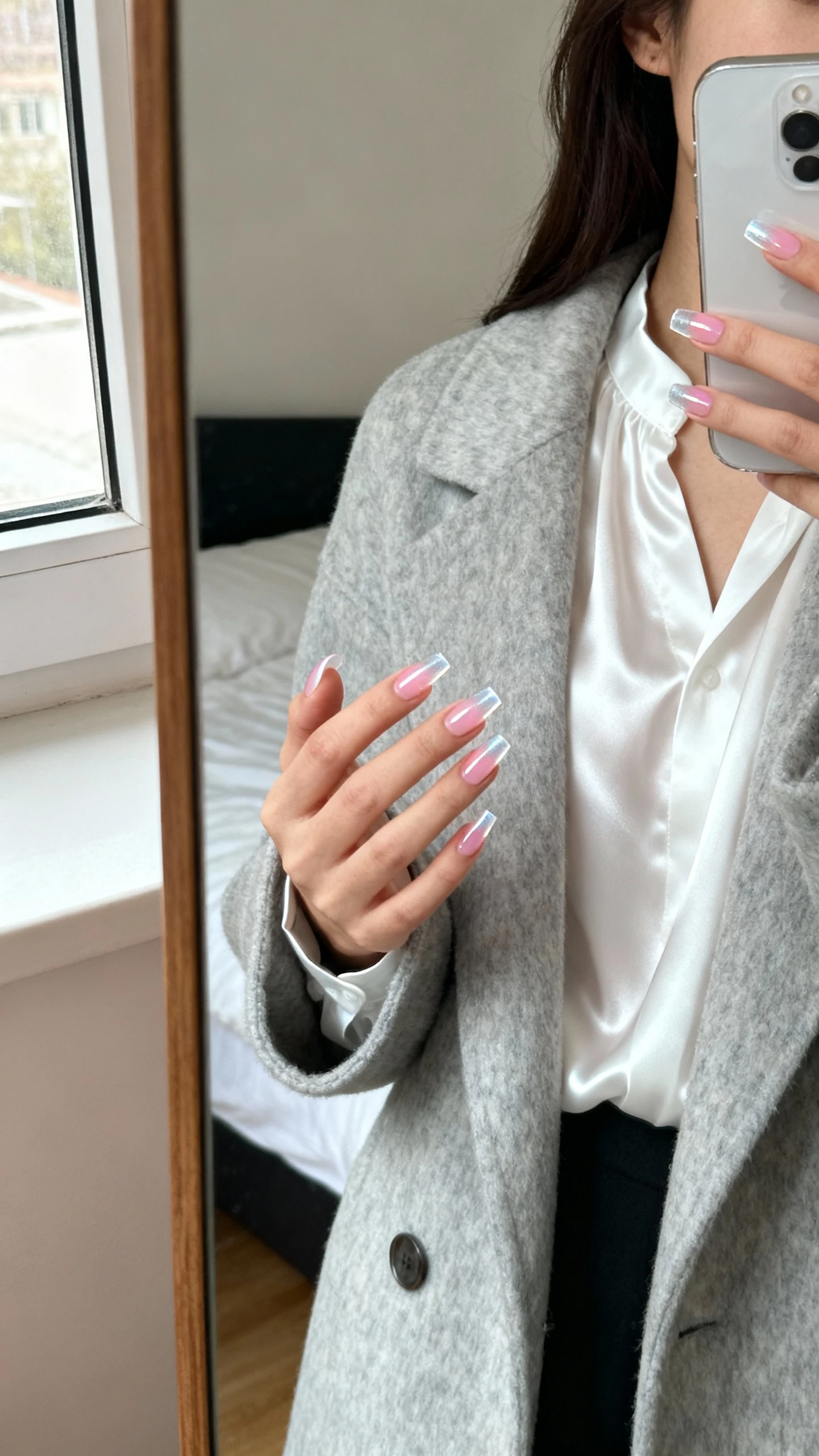 iPhone mirror selfie side angle of a woman with iced French tips (sheer pink base, frosted white tips with a soft glow), wearing a white satin blouse and light gray wool coat, face not visible, bedroom mirror, natural window light, iPhone photo quality.