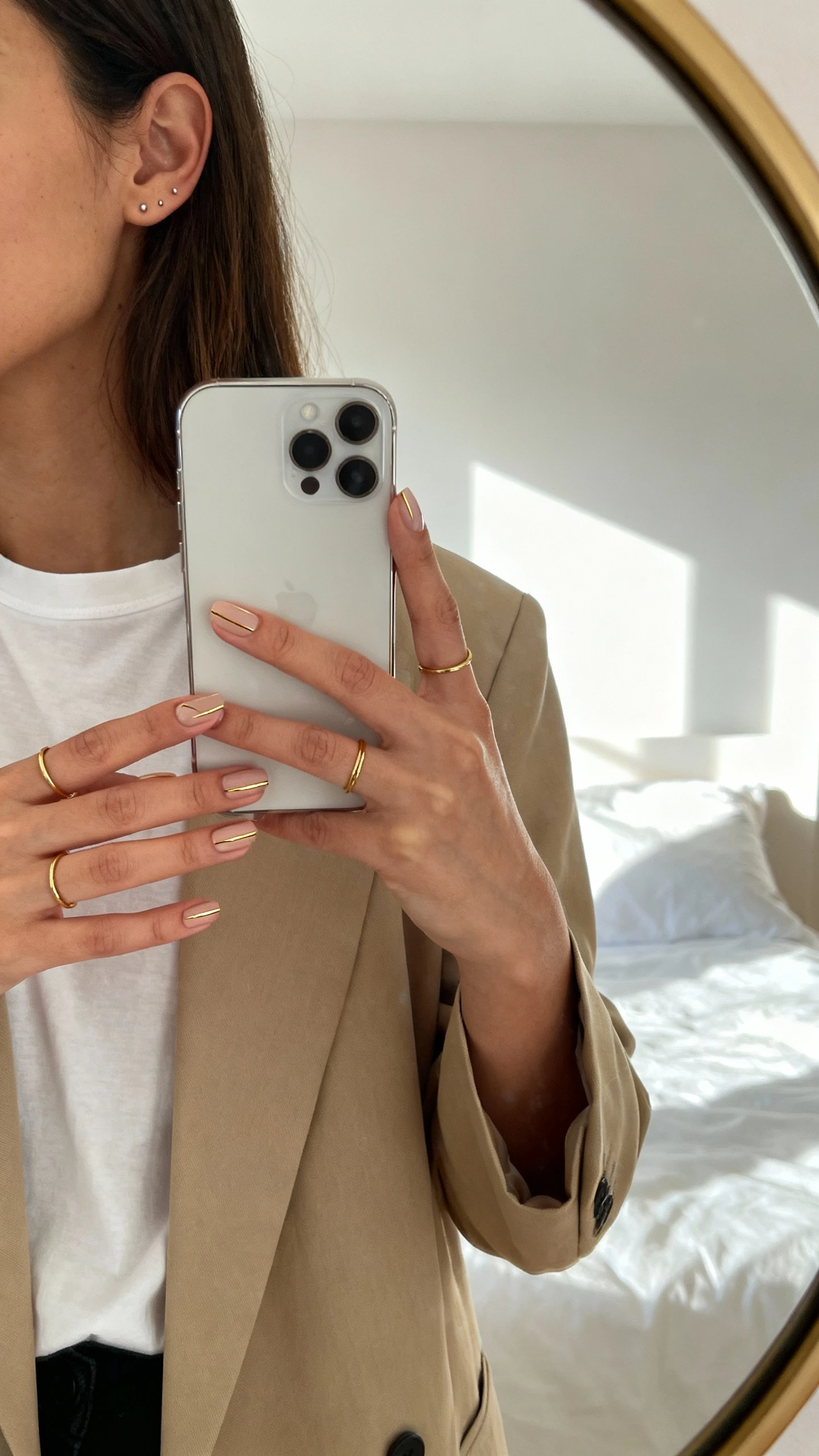 iPhone side-angle mirror selfie of a woman with gilded minimalism nails (thin metallic gold lines over sheer negative space), wearing a beige blazer, white tee, delicate gold rings, and minimalist studs, face not visible, bedroom mirror, morning window light, iPhone photo quality.