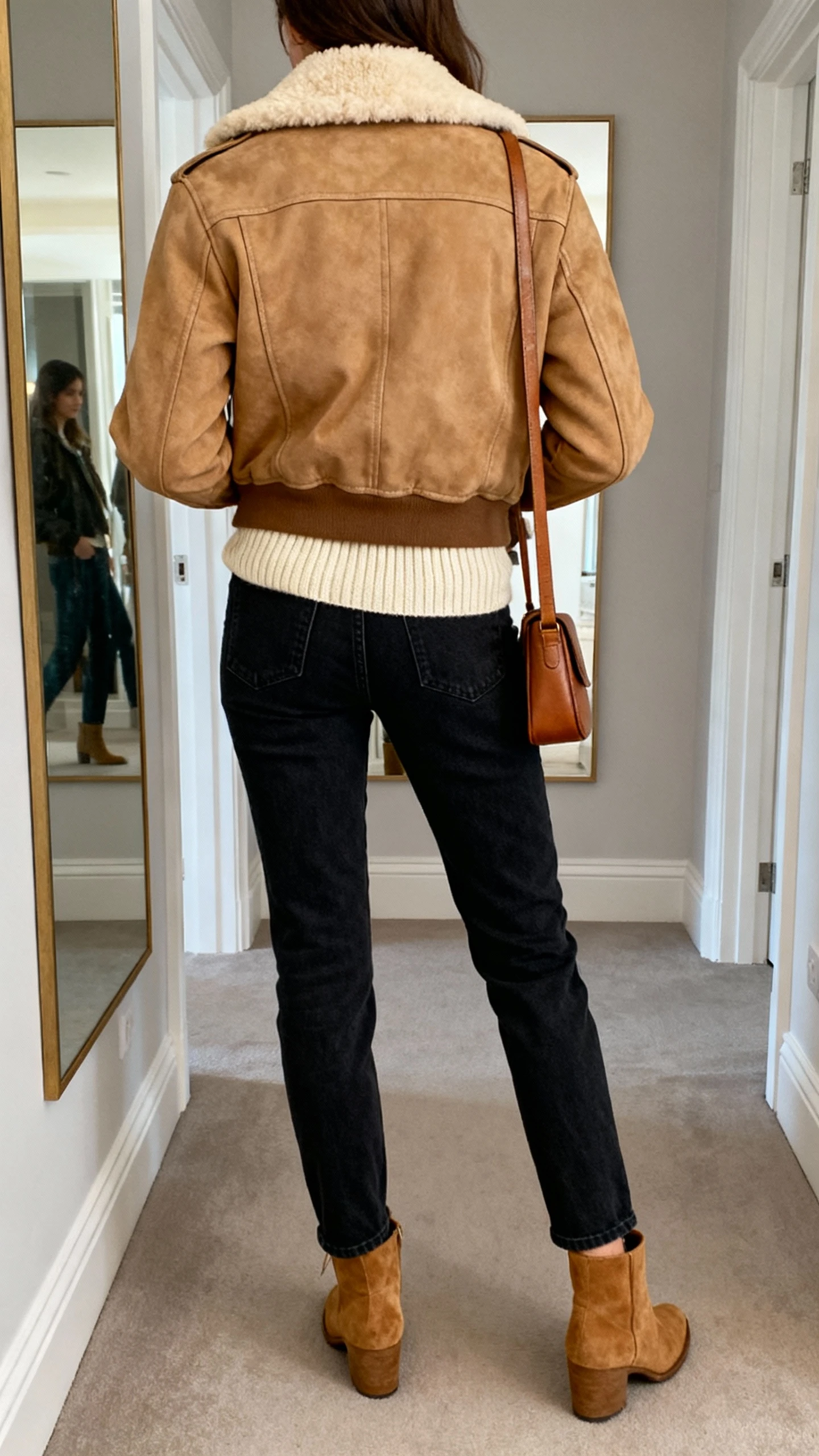 iPhone back-view photo of a woman wearing a tan shearling-collar aviator jacket over a cream ribbed knit, dark straight-leg jeans, tan suede ankle boots, and a simple leather crossbody, face not visible, hallway mirror, natural indoor light, iPhone photo quality.