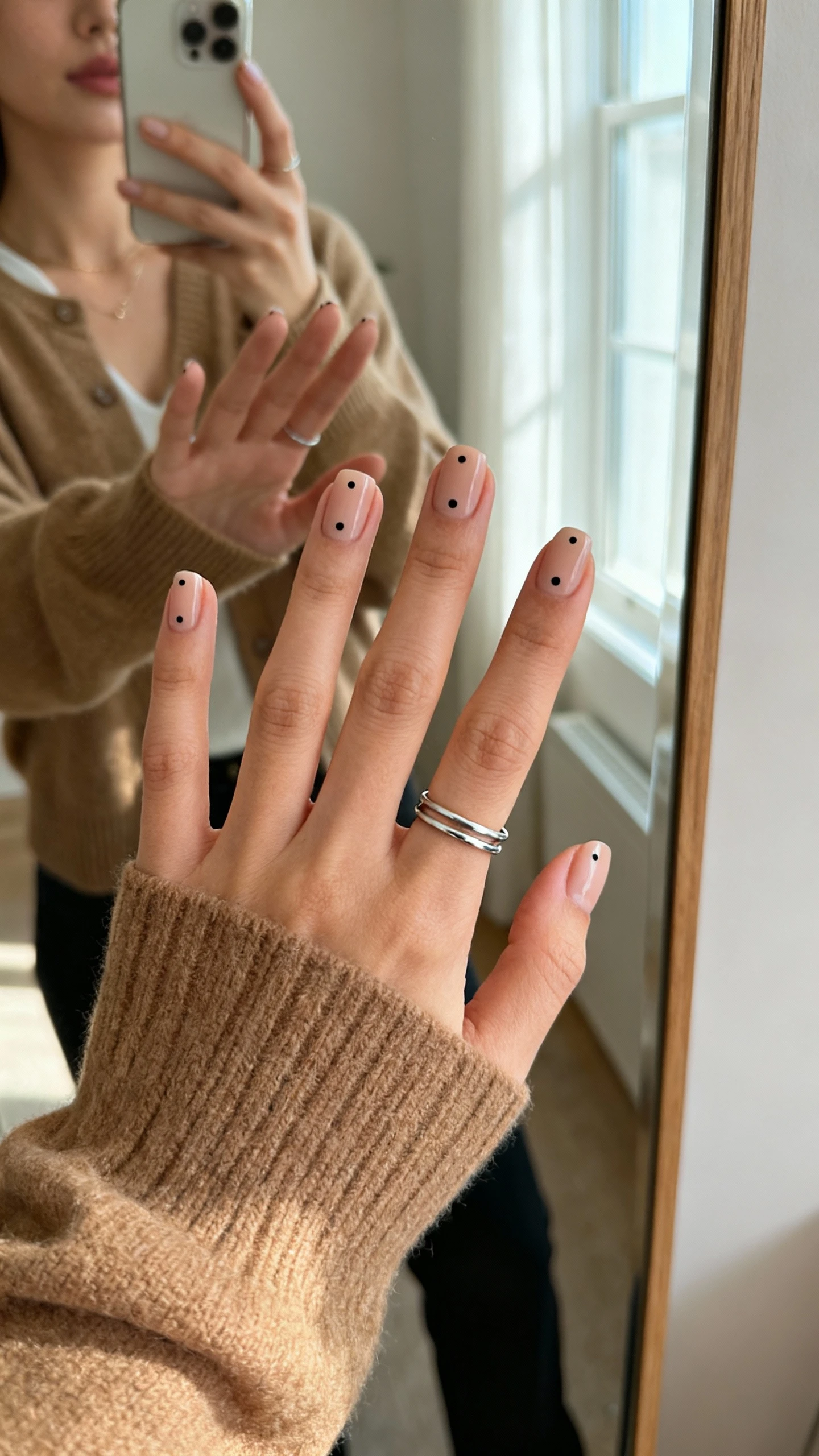 iPhone mirror selfie close-up of a woman’s hand with minimal dot accent nails (single tiny black dot near cuticle on each nail), wearing a tan cardigan and thin silver ring stack, face not visible, cropped at neck, full-length mirror, soft window light, iPhone photo quality