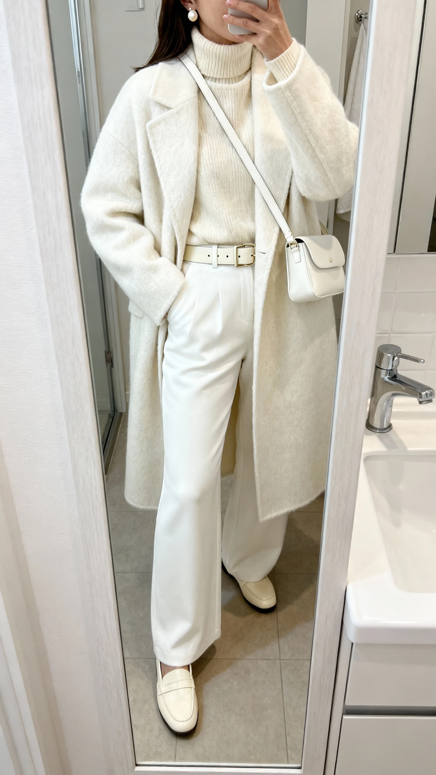 iPhone mirror selfie of a woman in a monochrome winter white look: ivory wool coat, cream cashmere turtleneck, white wide-leg trousers, off-white leather belt, cream loafers, pearl stud earrings, and a soft white crossbody, face not visible, bathroom full-length mirror, natural indoor lighting, iPhone photo quality.