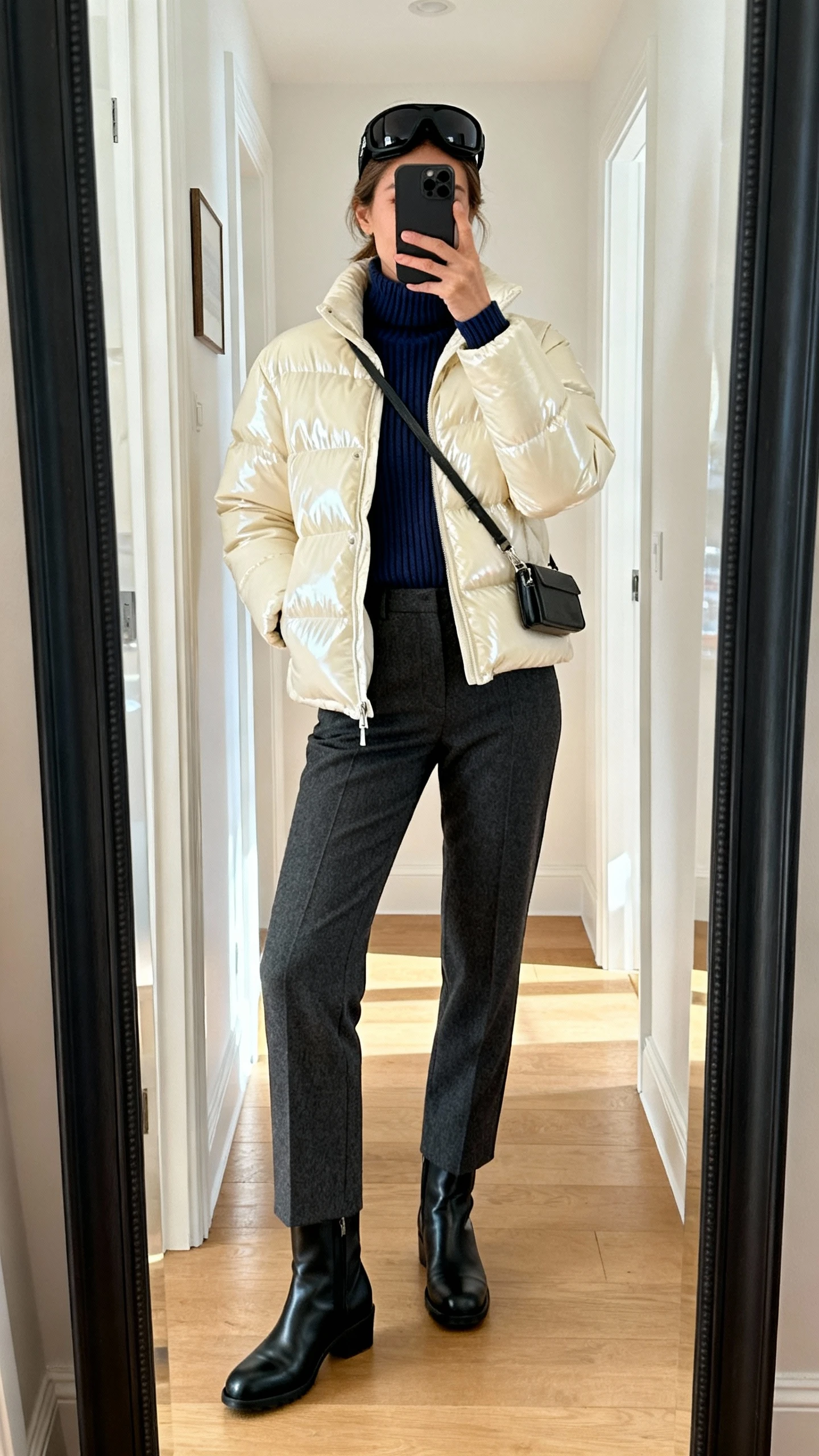iPhone mirror selfie of a woman in a ski-chic look: glossy cream puffer jacket over a navy fine-knit turtleneck, tailored wool trousers, black alpine-style sunglasses perched on head, black leather ankle boots, and a small crossbody, face not visible, entryway full-length mirror, bright daylight, iPhone photo quality.