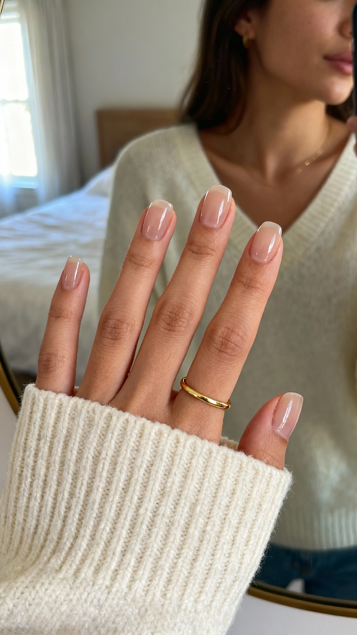 iPhone mirror selfie of a woman showing clean girl sheer gloss nails that go with everything, close-up hand over a cream sweater cuff and simple gold band ring, face not visible, cropped at neck, bedroom mirror, soft morning window light, iPhone photo quality