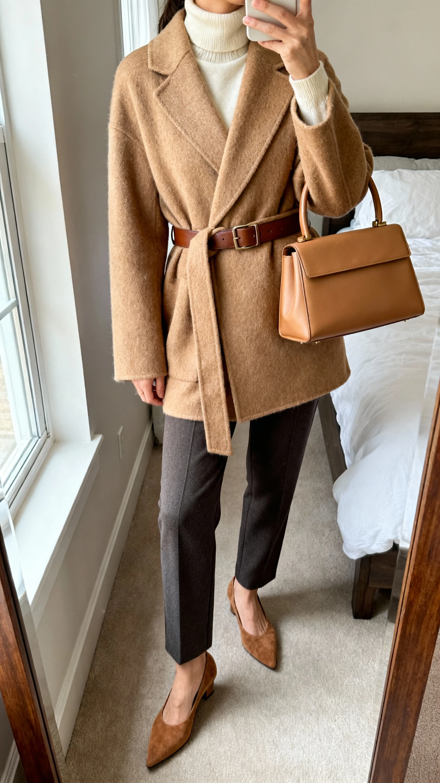 iPhone mirror selfie of a woman wearing a camel cashmere wrap coat over a cream turtleneck, tailored charcoal trousers, brown leather belt, camel suede pointed pumps, and a structured tan top-handle bag, face not visible, cropped at neck, bedroom mirror, morning window light, iPhone photo quality.