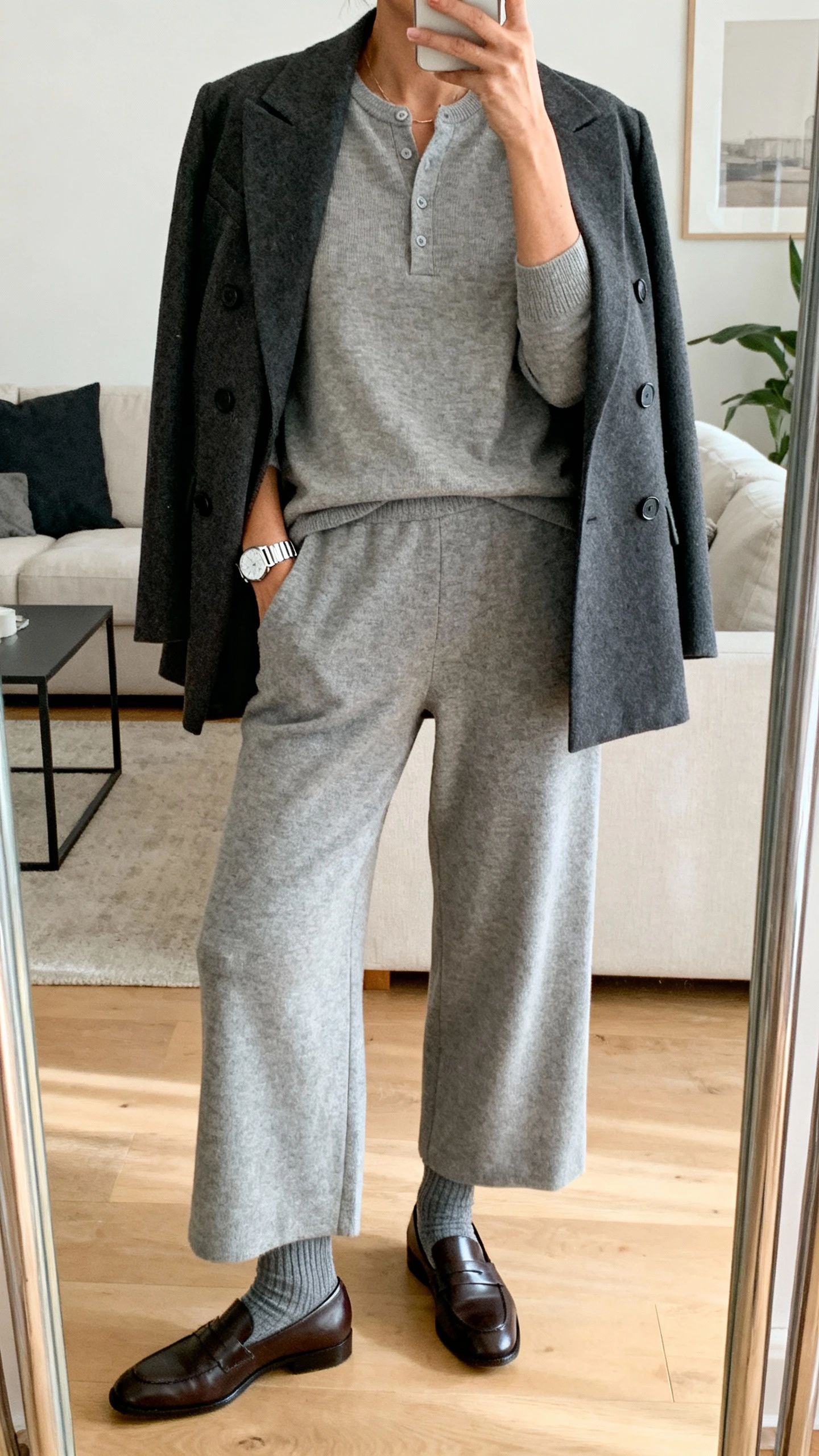 iPhone mirror selfie of a woman wearing a matching heather gray cashmere henley and wide-leg cashmere pants, draped with a charcoal double-breasted blazer, gray wool socks in sleek leather loafers, and a slim silver watch, face not visible, cropped at neck, living room mirror, natural morning light, iPhone photo quality.
