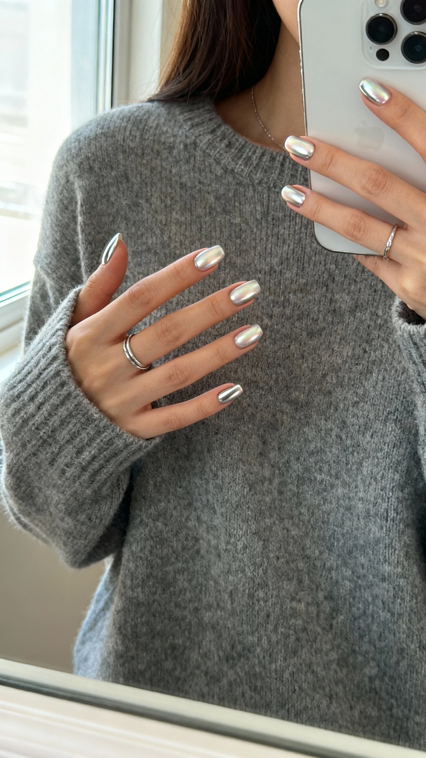 iPhone mirror selfie of a woman with soft chrome sheen nails (subtle pearly finish), wearing a gray knit sweater and dainty rings, face not visible, cropped at neck, near window light for gentle reflection, iPhone photo quality