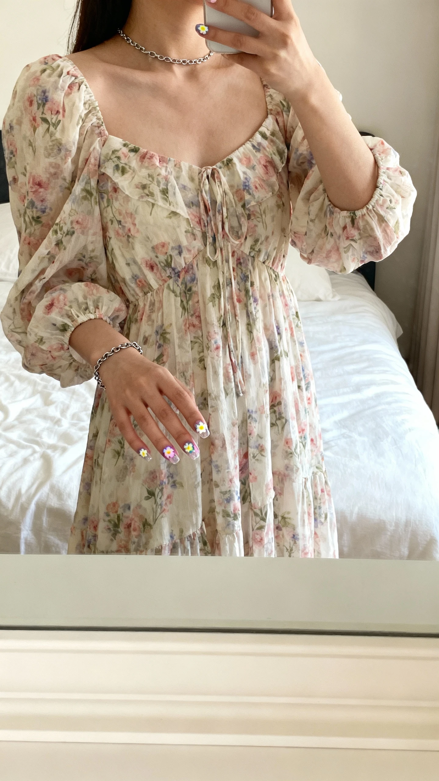 iPhone mirror selfie of a woman with tiny flower sticker nails that look hand-painted (small pastel floral decals on clear base), wearing a flowy floral dress and simple chain bracelet, face not visible, cropped at shoulders, bedroom mirror, soft morning light, iPhone photo quality